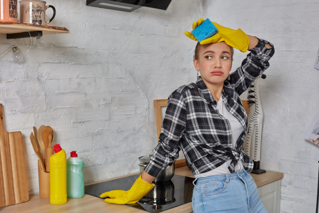 Woman in a kitchen wearing yellow cleaning gloves, holding a blue sponge to her forehead and looking frustrated while leaning against the counter near cleaning supplies and a stovetop.