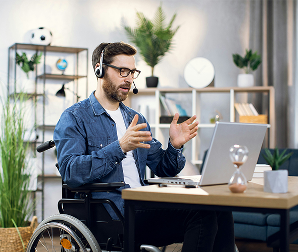 A man with glasses and a headset sits in a wheelchair at a wooden desk, gesturing with his hands while looking at a laptop screen. He is wearing a denim shirt over a white t-shirt. The background shows a bright, modern home office with potted plants and shelves.