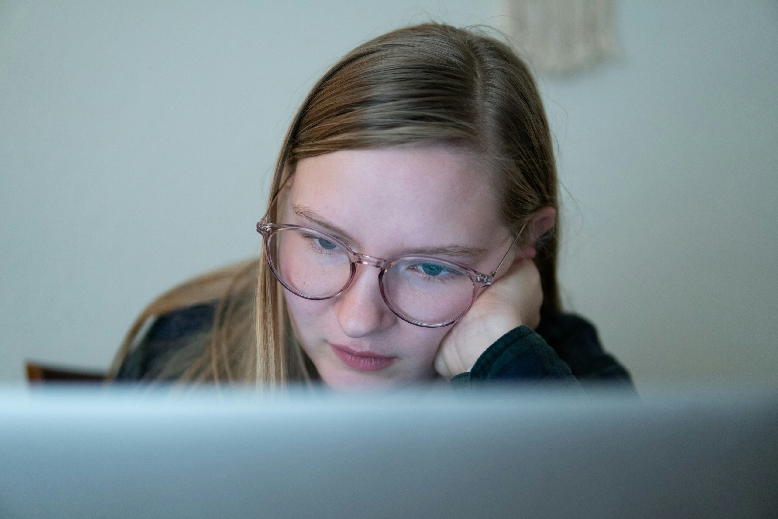 Young woman with glasses resting her head on her hand while looking intently at a laptop screen, appearing focused and thoughtful in a softly lit indoor setting.