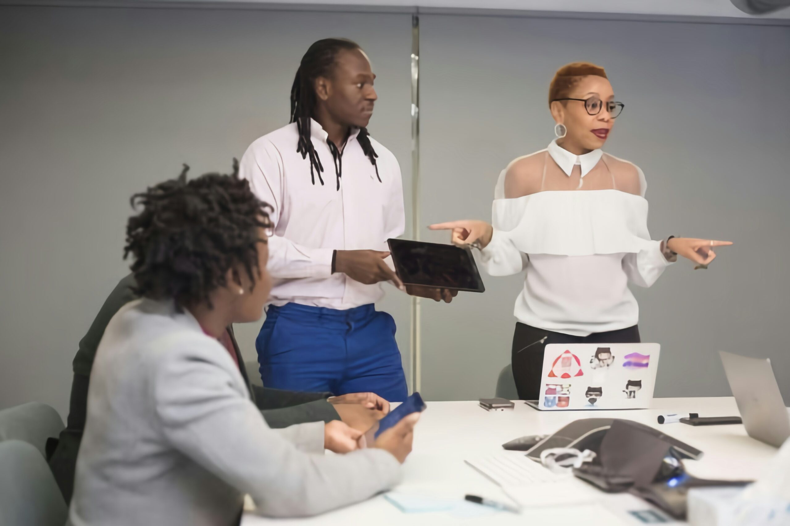 Two professionals stand at the front of a meeting room during a team discussion, with one holding a tablet and the other speaking and gesturing while seated colleagues listen around a conference table with laptops and phones.