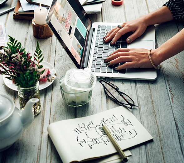 woman's hands typing on laptop on wooden table with a handwritten diagram next to laptop