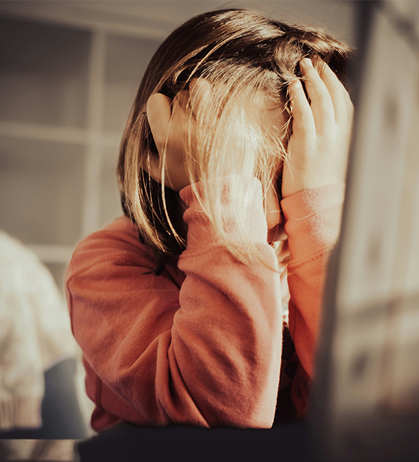 Person in an orange sweater sits at a desk with their head in their hands, appearing overwhelmed or stressed while looking toward a computer screen. Warm sunlight falls across their hair and face, creating a soft indoor scene.